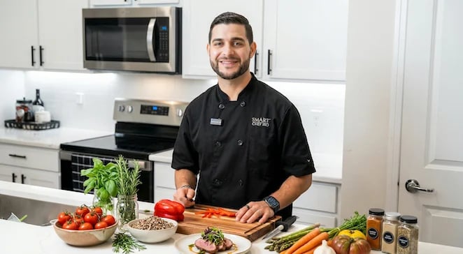 Man in black chefs coat smiling while chopping vegetables in a modern kitchen with fresh produce on counter